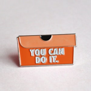 Op de afbeelding: Oranje emaille pin in de vorm van een doos, met een lichtoranje deksel. De pin heeft de woorden "YOU CAN DO IT." in het wit. Een motiverend accessoire.
