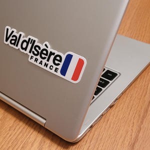 Könnte beinhalten: Ein silberner Laptop mit einem schwarz-weißen Aufkleber, auf dem "Val d'Isère France" und eine französische Flagge stehen.