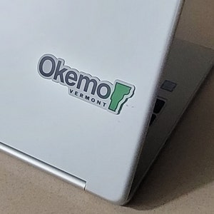 May include: A white laptop with a sticker that says "Okemo Vermont" and features a green outline of the state of Vermont.