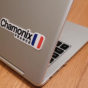 Op de afbeelding: Een zilveren laptop met een zwart-witte sticker waarop "Chamonix France" staat met een Frans vlagontwerp.