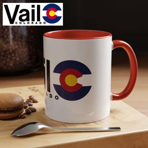 May include: White ceramic coffee mug with a red rim and handle. The mug has a black and blue graphic of the Colorado state flag with the word "Vail" and "Colorado" printed below the flag.