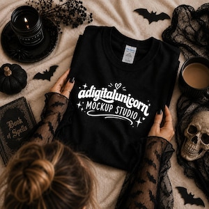 May include: A black t-shirt featuring white text that reads adigitalunicorn MOCKUP STUDIO with small star and heart accents. The shirt has a Gildan brand label at the inner neckline and is displayed on a light-colored fabric surface.