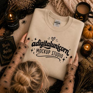 Könnte beinhalten: Ein beigefarbenes Sweatshirt mit Rundhalsausschnitt und schwarzem Schriftzug adigitalunicorn MOCKUP STUDIO, verziert mit kleinen Sternen und einem Herz. Das Kleidungsstück hat ein sichtbares Gildan-Markenetikett am inneren Halsausschnitt.