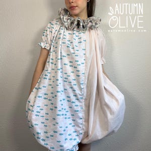 May include: A short-sleeved dress featuring a ruffled collar and a split design. One side displays a repeating pattern of small teal and light pink fish on a white background, while the other side is solid light pink. Text reads AUTUMN OLIVE.
