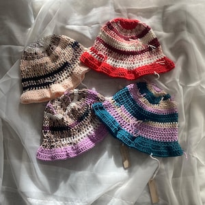 May include: Four handmade crochet bucket hats in various colors. One hat is red and white striped, another is blue and purple, and the other two are neutral tones with pink and black accents. Each hat has a frayed brim.