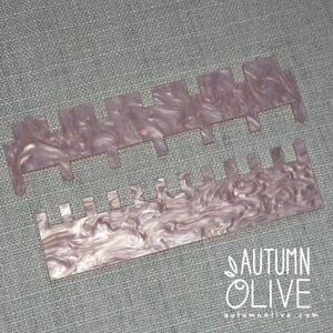 May include: Two rectangular tools made of a light purple, pearlescent material with a marbled pattern. Each tool features a series of evenly spaced, protruding rectangular teeth along one edge, with one tool having wider teeth than the other.