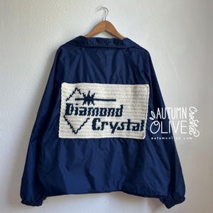 May include: A navy blue jacket with a cream-colored, crocheted patch on the back. The patch features the words "Diamond Crystal" in navy blue, with a geometric design above. The jacket is hanging on a wooden hanger.