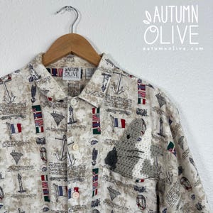 May include: A beige button-down shirt featuring a nautical print of lighthouses, anchors, and international flags with text. A gray and white crocheted creature is tucked into the chest pocket. The shirt has a collar and a brand label reading AUTUMN OLIVE.