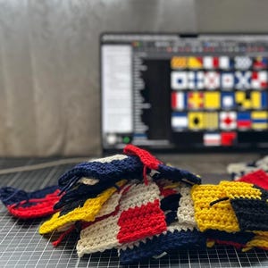 May include: Close-up of colourful, crocheted nautical signal flags in red, white, yellow, and navy blue. The flags are in various stages of completion, with a laptop displaying a digital chart of signal flags in the background. The flags are on a grey cutting mat.
