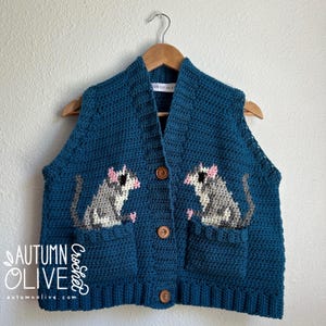 May include: A teal blue crochet vest with two pockets and a button-up front. The vest features two appliqued opossums with pink ears and paws. The text "Autumn Olive Crochet" is on the left side of the vest.