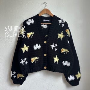 May include: A black, handmade crochet cardigan with a V-neck and wooden buttons. The sweater features white, yellow, and tan star and cloud motifs. The brand name "Autumn Olive" is visible in the upper left corner.