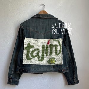 May include: A denim jacket with a cream-colored crochet patch featuring the word "tajin" in green, a red accent, and a lime graphic. The jacket is hanging on a wooden hanger against a white wall.