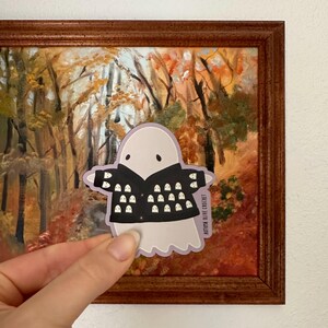 May include: A white ghost wearing a black jacket with a pattern of white ghosts. The text "AUTUMN OLIVE CROCHET" is printed on the bottom right corner of the sticker.