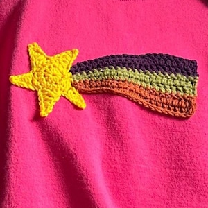 Puede incluir: Una sudadera rosa brillante con una aplicación de ganchillo de una estrella amarilla con una estela. La estela está compuesta por franjas horizontales de hilo morado oscuro, verde claro y naranja cosidas sobre la tela.