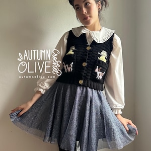 May include: Black crochet vest with a button-up front, featuring embroidered designs. The vest is worn over a white blouse with a ruffled collar and a blue tulle skirt. The text "AUTUMN OLIVE" is visible on the left side of the image.