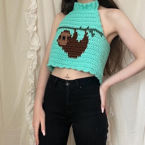 May include: A turquoise crochet halter top with a brown sloth design. The top has a high neck and a scalloped edge. The sloth is hanging from a branch. The top is handmade and appears to be a summer garment.