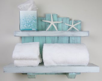 May include: Two-tiered wooden shelf with a distressed light blue paint finish. The top shelf has a box of tissues and two white starfish decorations. The bottom shelf has two white towels folded on it.