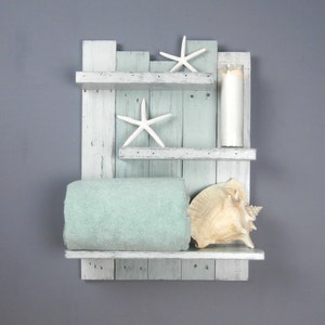 May include: A rustic, wall-mounted shelf unit with a coastal theme. The unit is made of distressed, light blue and white painted wood. It features two shelves, a rolled aqua towel, two white starfish, a seashell, and a tall candle.