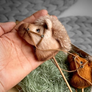 Könnte beinhalten: Nahaufnahme von zwei Miniatur-Pferdeköpfen aus Plüsch, beige und braun, mit detaillierten Zaumzeugen. Auf Holzstäben montiert, wirken sie wie ein handgefertigtes Spielzeug. Der Hintergrund ist verschwommen, mit Grün- und Grautönen.