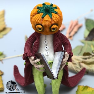 May include: A crocheted pumpkin-headed figure wearing a burgundy coat and green pants holds a closed book. The figure has large eyes and a green stem on top of its head.