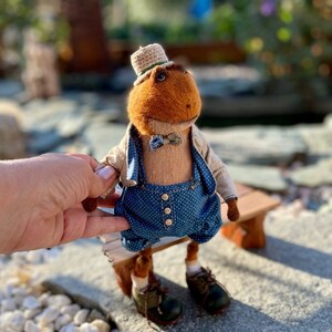 May include: A brown plush frog wearing a blue and white overalls, a brown bow tie, a straw hat, and brown shoes. The frog is sitting on a wooden bench.