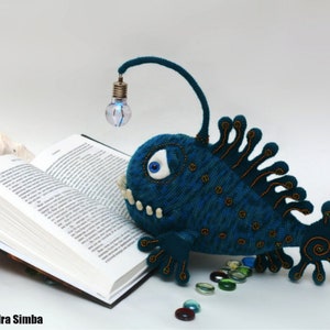 May include: A blue crocheted anglerfish with a light bulb on top, reading a book. The fish has a large eye and a row of teeth. There are small glass beads scattered around the fish.
