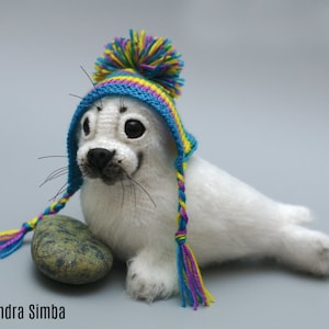 Puede incluir: Un juguete de peluche de foca blanca que lleva un gorro de punto colorido con un pompón y trenzas. El gorro presenta rayas azules, amarillas y moradas. Una pequeña piedra verde descansa junto a la foca. El texto "Alexandra Simba" está en la parte inferior izquierda.