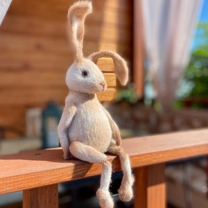 May include: A beige knitted bunny rabbit toy with long ears sits on a wooden railing. The rabbit has a white belly and brown eyes.