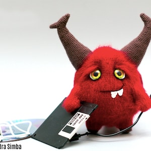 May include: A red crocheted monster with horns and a toothy grin holds a black floppy disk and a USB cable. The monster has yellow eyes and a white smile.