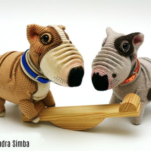 Puede incluir: Dos bulldogs de peluche tejidos a crochet, uno marrón y otro gris, están jugando en un sube y baja de madera. El bulldog marrón tiene un collar azul y el bulldog gris tiene un collar naranja.