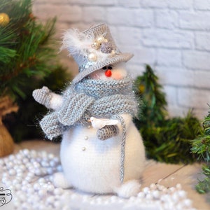 Könnte beinhalten: Ein weißer gehäkelter Schneemann, der einen grauen Hut mit einer weißen Feder, einen grauen Schal mit silbernem Glitzer und weiße Handschuhe trägt. Der Schneemann hält einen weißen gehäkelten Vogel in seiner rechten Hand und einen grauen gehäkelten Stock in seiner linken Hand. Der Schneemann sitzt auf einer weißen Oberfläche mit weißen Perlen, die um ihn herum verstreut sind.