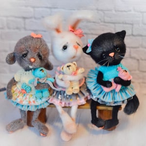 Puede incluir: Tres animales de peluche tejidos a crochet sentados en un banco de madera. Un ratón gris con un lazo rosa sostiene un animal de peluche azul. Un conejito blanco con un lazo rosa sostiene un osito de peluche marrón. Un gato negro con un lazo rosa sostiene un animal de peluche rosa.