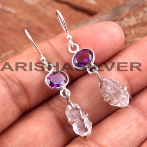 May include: A pair of silver earrings with a purple amethyst gemstone and a clear quartz crystal. The earrings are dangle style and have a hook closure.