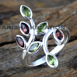 May include: A silver ring with a branch design featuring green and red gemstones. The ring is set on a wooden surface.