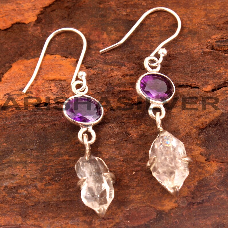 May include: A pair of silver dangle earrings with a purple amethyst gemstone and a clear quartz crystal drop.