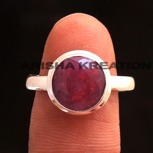 May include: A silver ring with a large, round, faceted ruby gemstone. The ruby is a deep red color and is set in a bezel setting. The ring is shown on a finger. The text "ARISHA KREATION" is visible.