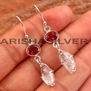 May include: Silver dangle earrings with a red gemstone and a clear quartz crystal. The earrings have a hook closure.