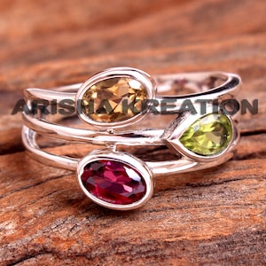 May include: Silver ring featuring three gemstones: a pear-shaped green stone, a round yellow stone, and a round red stone.