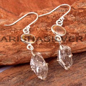 May include: Silver earrings with a clear quartz crystal and a yellow oval gemstone. The earrings have a hook closure.