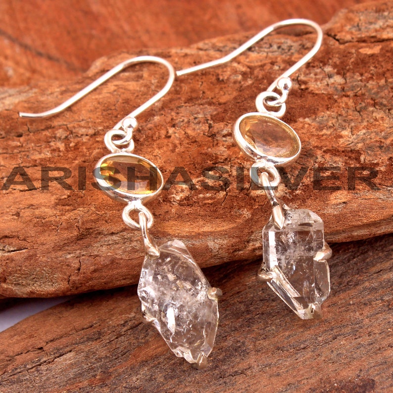 May include: Silver earrings with a clear quartz crystal and a yellow oval gemstone. The earrings have a hook closure.