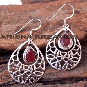May include: A pair of silver dangle earrings with a red gemstone in the center of each earring. The earrings have a delicate, filigree design.