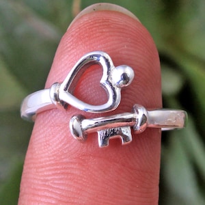 Puede incluir: Un anillo de plata con un diseño de llave en forma de corazón. El anillo se lleva en un dedo.