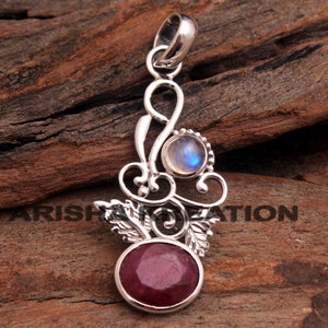 Puede incluir: Un colgante de plata con un rubí rojo y una piedra de luna blanca. El colgante tiene forma de diseño floral con hojas y espirales.