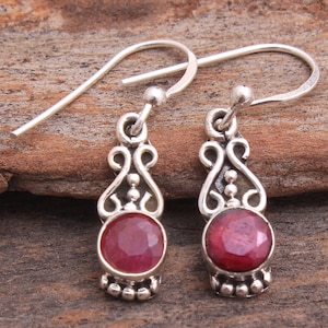 May include: A pair of silver dangle earrings with red gemstone accents. The earrings feature a delicate, ornate design with a round, faceted gemstone in the centre.