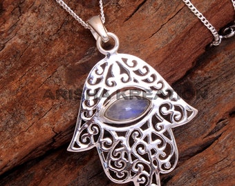 Könnte beinhalten: Silberner Hamsa-Hand-Anhänger mit einem blauen Mondstein in der Mitte. Der Anhänger ist aufwendig mit einem filigranen Design verziert.