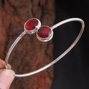 Könnte beinhalten: Ein silbernes Armreif-Armband mit zwei roten Edelstein-Akzenten. Das Armband ist offen und hat ein schlichtes Design.