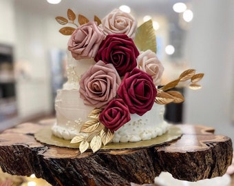 Può includere: Una torta nuziale bianca decorata con rose rosa e bordeaux e dettagli in foglia d'oro. La torta è esposta su un supporto per torte in legno rustico.