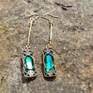 Puede incluir: Un par de pendientes colgantes dorados. Cada pendiente presenta una barra larga y delgada de oro conectada a un colgante rectangular. El colgante tiene una gran gema verde esmeralda rodeada por un diseño de filigrana negro y dorado. Los pendientes se muestran sobre una superficie de piedra gris texturizada.