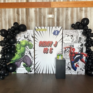 May include: A birthday party backdrop with a comic book theme. The backdrop features images of Hulk, Spiderman, and a birthday message that says "Remy J is 5". Black balloons are arranged on either side of the backdrop.