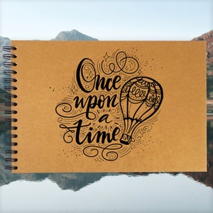 Può includere: Un diario in carta kraft marrone con rilegatura a spirale. La copertina presenta un'illustrazione in bianco e nero di una mongolfiera con il testo "Once upon a time" in un carattere decorativo.